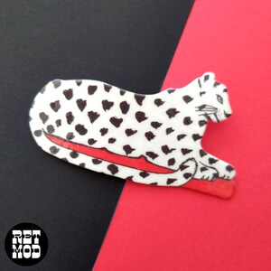 May include: A white and black spotted leopard brooch with a red base. The brooch is lying on a red and black background. The brooch has a retro mod design.