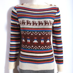 May include: A vintage-style sweater with a cream-colored neckline and cuffs, adorned with brown buttons on the shoulder. The sweater features horizontal stripes in various colors, including blue, red, and brown, and includes patterned sections with llama and figure motifs.