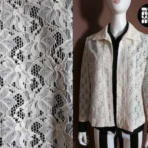 May include: Cream-colored lace jacket with long sleeves and a collared neckline. The jacket is open in the front, revealing a black shirt underneath. The lace pattern features floral designs. The mannequin is wearing black and white striped pants.