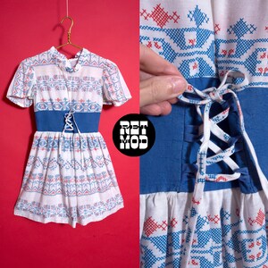 May include: A vintage white dress with short sleeves, featuring a blue and red embroidered pattern. The dress has a blue corset-style waist and a lace-up detail. The dress is hanging on a gold hanger against a red background.
