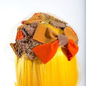 Puede incluir: Un sombrero vintage con velo de red y lazos de tela coloridos. Los lazos son en tonos naranja, marrón y beige. El sombrero se coloca sobre cabello amarillo, creando un contraste llamativo. Accesorio ideal para un look retro o de disfraz.