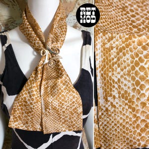 Puede incluir: Una bufanda con estampado de piel de serpiente en beige y blanco está drapeada alrededor del cuello de un maniquí, asegurada con un cierre de metal plateado. El maniquí lleva una parte superior con estampado en blanco y negro. La imagen incluye un primer plano de la textura de la bufanda y el logo "RET MOD".