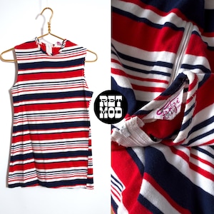 May include: A sleeveless dress with horizontal stripes in red, white, and navy blue. The dress has a high neckline and a zip. The label says "Fritzi of California". The dress is hanging on a gold hanger.