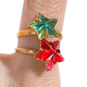 May include: Two gold-toned metal rings featuring star-shaped faceted stones. The top ring has a translucent green star, while the bottom ring features a vibrant red star. Both bands are thin and metallic, showcasing a simple, classic design.
