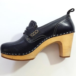 May include: A navy blue leather clog shoe with a light wood heel and sole. The shoe features white stitching, a strap across the top, and a small loop at the back. The heel measures approximately 3 inches.
