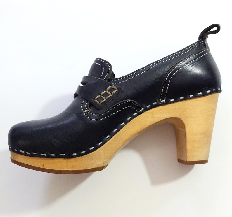 May include: A navy blue leather clog shoe with a light wood heel and sole. The shoe features white stitching, a strap across the top, and a small loop at the back. The heel measures approximately 3 inches.