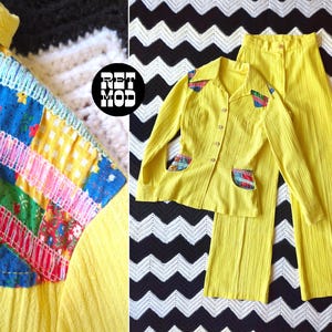 May include: A vintage yellow two-piece suit. The long-sleeved shirt has patchwork detailing on the pockets and shoulders. The matching trousers complete the set. The outfit is displayed on a black and white patterned background.