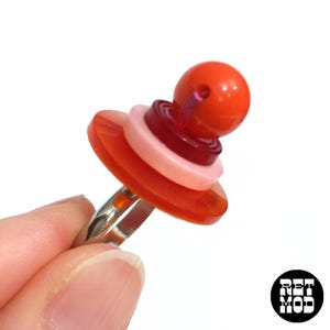 May include: A ring with a stack of red, pink, and orange buttons. The top button is a sphere.