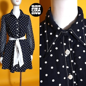 May include: Navy blue polka dot dress with sheer long sleeves and a white tie belt. The dress features white polka dots and a button-down front. The Retro Mod logo is visible in the upper left corner.