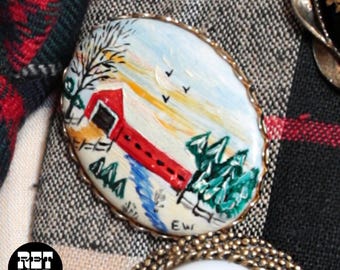 May include: Oval-shaped brooch with a hand-painted landscape scene. The artwork depicts a red covered bridge, trees, and a blue sky with birds. The brooch has a gold-colored, textured border and is set against a plaid background.