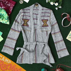 May include: A gray knit cardigan featuring long sleeves, an open front with a matching tie belt, and horizontal purple and gold stripes. The upper chest area displays two symmetrical geometric shapes in green, gold, and purple thread accents.