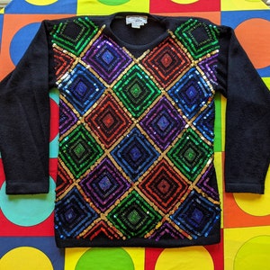 Pode incluir: Camisola preta com um padrão geométrico colorido de lantejoulas. As lantejoulas estão dispostas em forma de diamante e são em tons de vermelho, verde, azul, roxo e dourado.