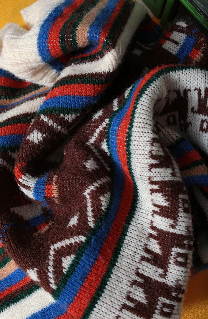 May include: Close-up of a folded, patterned sweater. The sweater features a repeating geometric design in brown, white, blue, red, and green. The knitwear has a textured appearance, with a cream-colored collar.