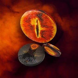 Può includere: Tre gettoni circolari con l'immagine di un occhio ardente con pupilla verticale, una fortezza scura sormontata da un occhio luminoso e un anello dorato. I gettoni presentano colori vivaci arancione, rosso, giallo e nero con finitura lucida.