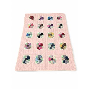 May include: A rectangular quilt featuring a solid light pink background with a grid of twenty square patches. Each patch contains a circular floral motif made from various patterned fabric scraps in shades of blue, purple, red, and green.