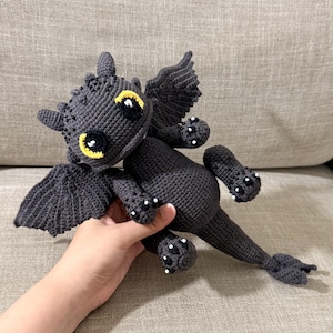 May include: A handmade black crochet dragon with yellow eyes and white accents. The dragon has wings, four legs, and a long tail. The dragon is lying on a gray couch.