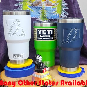 May include: Three 30-ounce YETI Rambler tumblers with different state outlines and the iconic lightning bolt graphic. The tumblers are white, green, and blue. The tumblers are on a wooden surface with yellow and blue coasters.