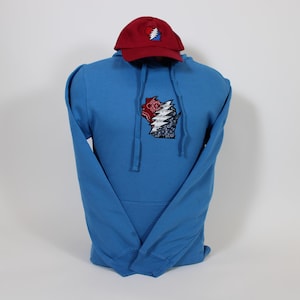 May include: A blue hooded sweatshirt with a Grateful Dead inspired graphic of a rose and lightning bolt on the front. A red baseball cap with a Grateful Dead logo is on top of the sweatshirt.