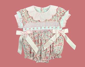 May include: A pink and white baby onesie with a floral print and pink bows. The onesie has a scalloped collar and short sleeves.