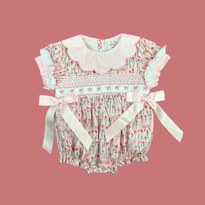 Peut inclure: Une barboteuse rose et blanche pour b&eacute;b&eacute; avec un imprim&eacute; floral et des n&oelig;uds roses. La barboteuse a un col festonn&eacute; et des manches courtes.