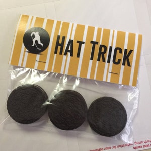 May include: A clear plastic bag with a yellow and white striped label that says "HAT TRICK". The label has a black circle with a white silhouette of a hockey player. There are three chocolate cookies inside the bag.