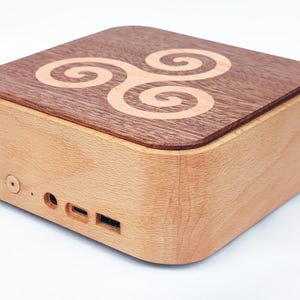 Può includere: Scatola di legno quadrata con angoli arrotondati. Il coperchio è in legno più scuro con un motivo a spirale chiaro. I lati sono in legno più chiaro. Misura circa 15 cm di larghezza e ha diverse porte.