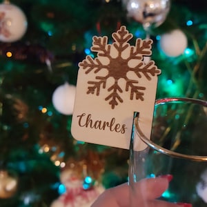 Peut inclure: Ornement de Noël en bois en forme de flocon de neige avec le nom "Charles" gravé. L'ornement est conçu pour être accroché à un verre. L'arrière-plan est un sapin de Noël flou.