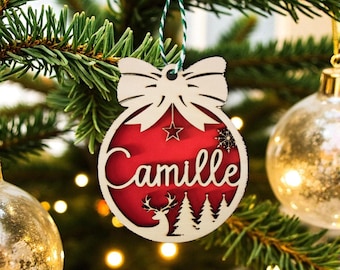 Peut inclure: Ornement de Noël en bois en forme de nœud papillon et de boule. La boule rouge porte le nom "Camille" en écriture blanche. Une étoile, un flocon de neige, un cerf et des arbres sont également découpés. Suspendu à une ficelle verte et blanche.
