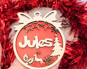 Peut inclure: Une décoration de Noël en bois avec le nom "Jules" en écriture cursive. L'ornement présente un fond rouge avec un cerf, un flocon de neige et un motif de sapin de Noël. L'ornement est entouré d'une guirlande de tinsel rouge.