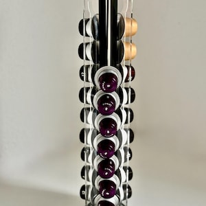 May include: A clear acrylic coffee pod holder with a black base and top. The holder is filled with various coffee pods in shades of black, gold, and purple. The word "COFFEE" is visible on the base.
