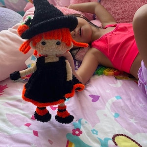 May include: A handmade crochet doll dressed as a witch. The doll has a black hat, black dress with orange trim, and orange hair. The doll is approximately 8 inches tall. The doll is on a pink patterned bedspread.