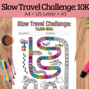 May include: A travel challenge printable with the text "Slow Travel Challenge: 10K" and "10,000 GOAL". The design includes a path with travel-related icons. A cup of coffee and colorful pens are on a wooden surface.