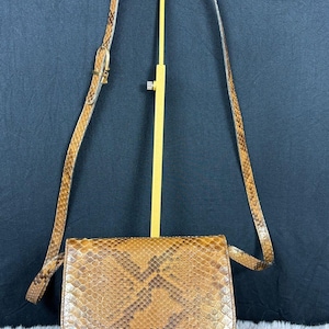 May include: A brown and tan snakeskin patterned crossbody bag with a rounded bottom edge and a long, adjustable shoulder strap. The bag features a textured, reptilian-inspired scale pattern throughout the exterior surface and a gold-toned metal buckle.