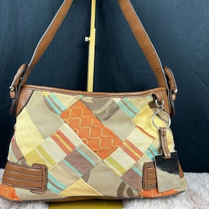 May include: A shoulder bag featuring a patchwork design with various fabric textures in shades of orange, yellow, brown, and teal. The bag has brown leather trim, a single handle, and a silver-toned metal rectangular charm hanging from a ring.