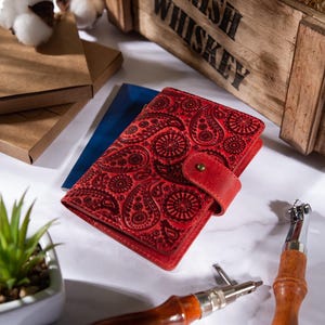May include: A red leather wallet with a paisley pattern and a snap closure. The wallet is open, revealing a blue document inside. The image also shows a wooden box, small brown boxes, and a small plant.