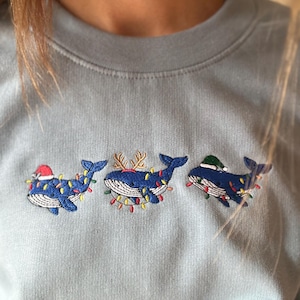 Pode incluir: Camisola azul claro com baleias bordadas decoradas com luzes de Natal e chapéus festivos. As baleias são adornadas com um gorro de Pai Natal, chifres de rena e um gorro de inverno verde.