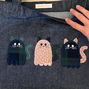 Peut inclure: Un sac fourre-tout en jean bleu foncé avec une étiquette blanche portant le texte Studio Cozy. Trois appliqués en forme de fantômes chats sont cousus sur le devant. Les chats sont en tissu à carreaux vert, à fleurs roses et à carreaux bleus.