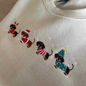 Peut inclure: Un sweat-shirt beige orné de broderies de teckels en tenues de Noël. Les chiens portent un serre-tête en forme de bois de renne, un bonnet de Père Noël, un pull rayé et un chapeau en forme de sapin de Noël. Des flocons de neige sont également brodés.