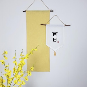 May include: Two wall hangings. One is a long, rectangular, yellow banner. The other is a white pennant with black characters and a tassel. Both are suspended by wooden rods and twine.