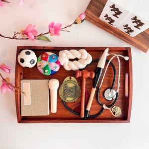 May include: A wooden tray filled with various objects, including a soccer ball, a colorful toy, a rope knot, a book, a microphone, a gavel, a stethoscope, and a brush. Pink flowers and decorative paper are also present.