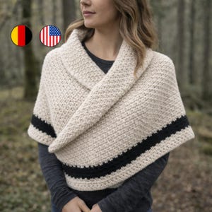 May include: A cream-coloured crocheted shawl featuring a wide collar and black stripes along the lower edge. The shawl is worn over a dark grey long-sleeved top. The background is a blurred forest scene.