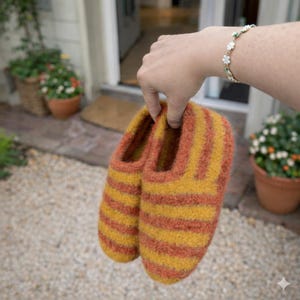 Può includere: Un paio di pantofole fatte a mano con un motivo a righe gialle e arancioni. Le pantofole sono tenute in mano, mostrando il loro aspetto morbido e strutturato. Le calzature sembrano essere fatte di lana o materiale simile.