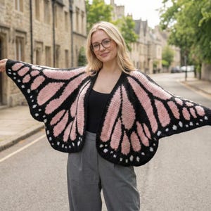 May include: A knitted butterfly wing-shaped cardigan in pink and black. The open-front design has a black outline with pink sections, and white dots. The cardigan is made of a soft, knitted material.