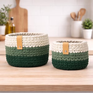 Puede incluir: Dos cestas de ganchillo hechas a mano en diferentes tamaños. Las cestas presentan un diseño de bloques de color con secciones crema, verdes y verde oscuro. Cada cesta tiene una correa de cuero marrón claro sujeta con un remache metálico. Las cestas están sobre una superficie de madera.