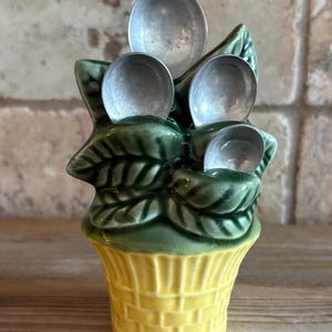 May include: A ceramic measuring spoon set featuring four silver-toned metal spoons nested within a green leafy plant structure. The plant is housed in a yellow, basket-weave textured ceramic pot with a visible crackle glaze finish throughout.