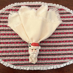 May include: An oval quilted placemat featuring a repeating pattern of red, green, and white stripes with a white ruffled border. A cream-colored fabric napkin with fringed edges is held together by a ceramic Santa Claus napkin ring.