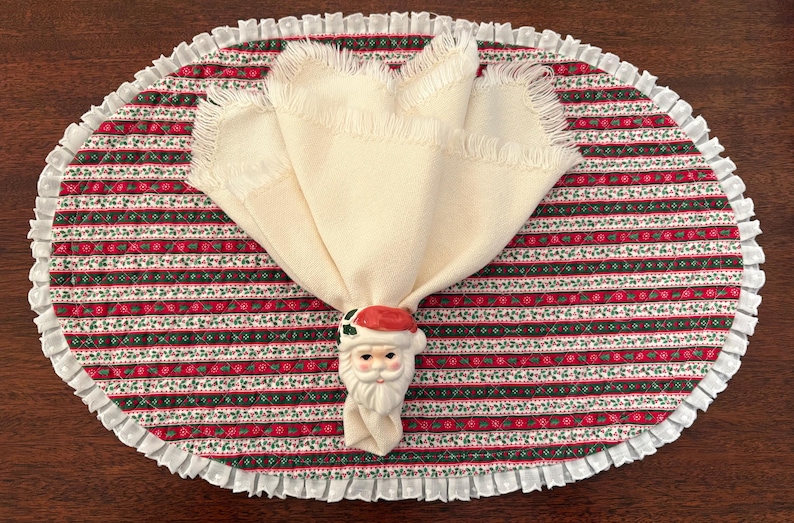 May include: An oval quilted placemat featuring a repeating pattern of red, green, and white stripes with a white ruffled border. A cream-colored fabric napkin with fringed edges is held together by a ceramic Santa Claus napkin ring.