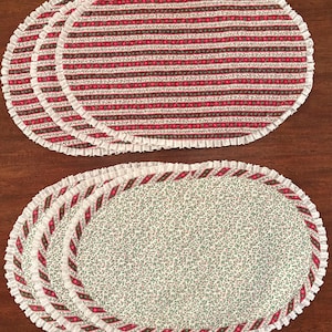 May include: A set of oval placemats featuring two distinct designs. One set displays horizontal stripes in red, green, and white, while the other features a small green floral pattern on a white background, both finished with a white ruffled edge.