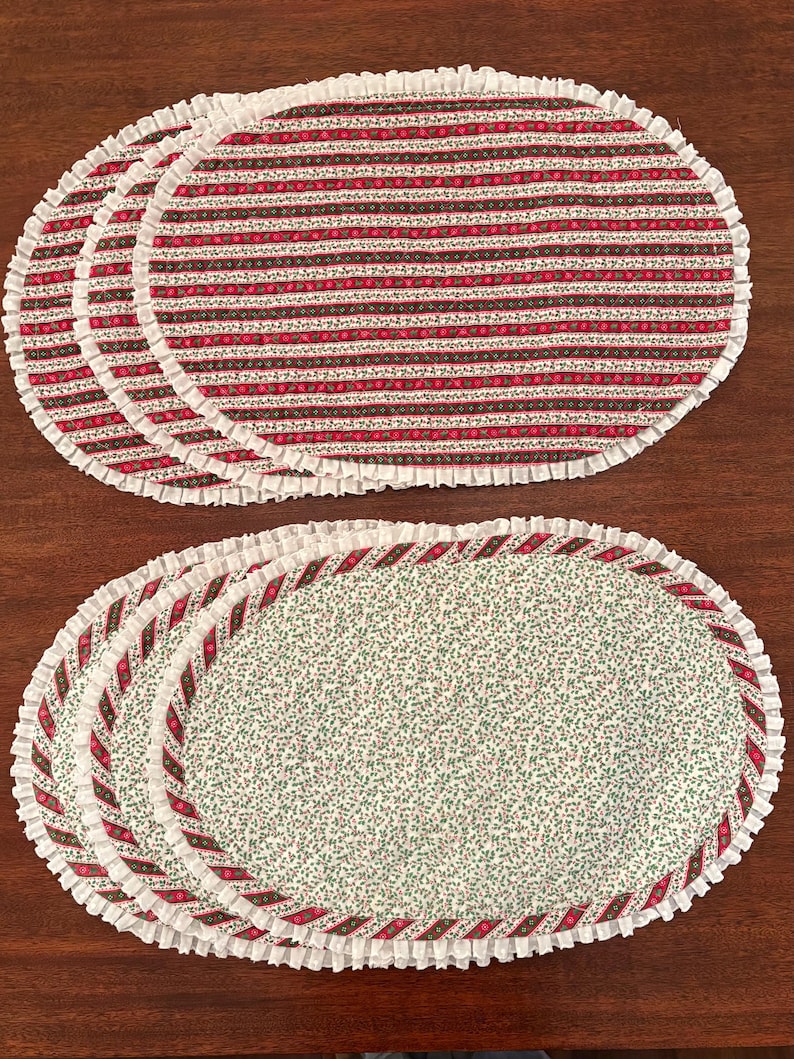 May include: A set of oval placemats featuring two distinct designs. One set displays horizontal stripes in red, green, and white, while the other features a small green floral pattern on a white background, both finished with a white ruffled edge.