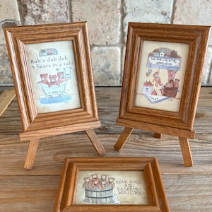 May include: Three framed pictures with brown wooden frames. Two are on small easels, and one is flat. The pictures feature teddy bears in various scenes, with text that reads "Rub a dub dub... 3 bears in a tub!" and "Bear hugs and backrubs welcome."